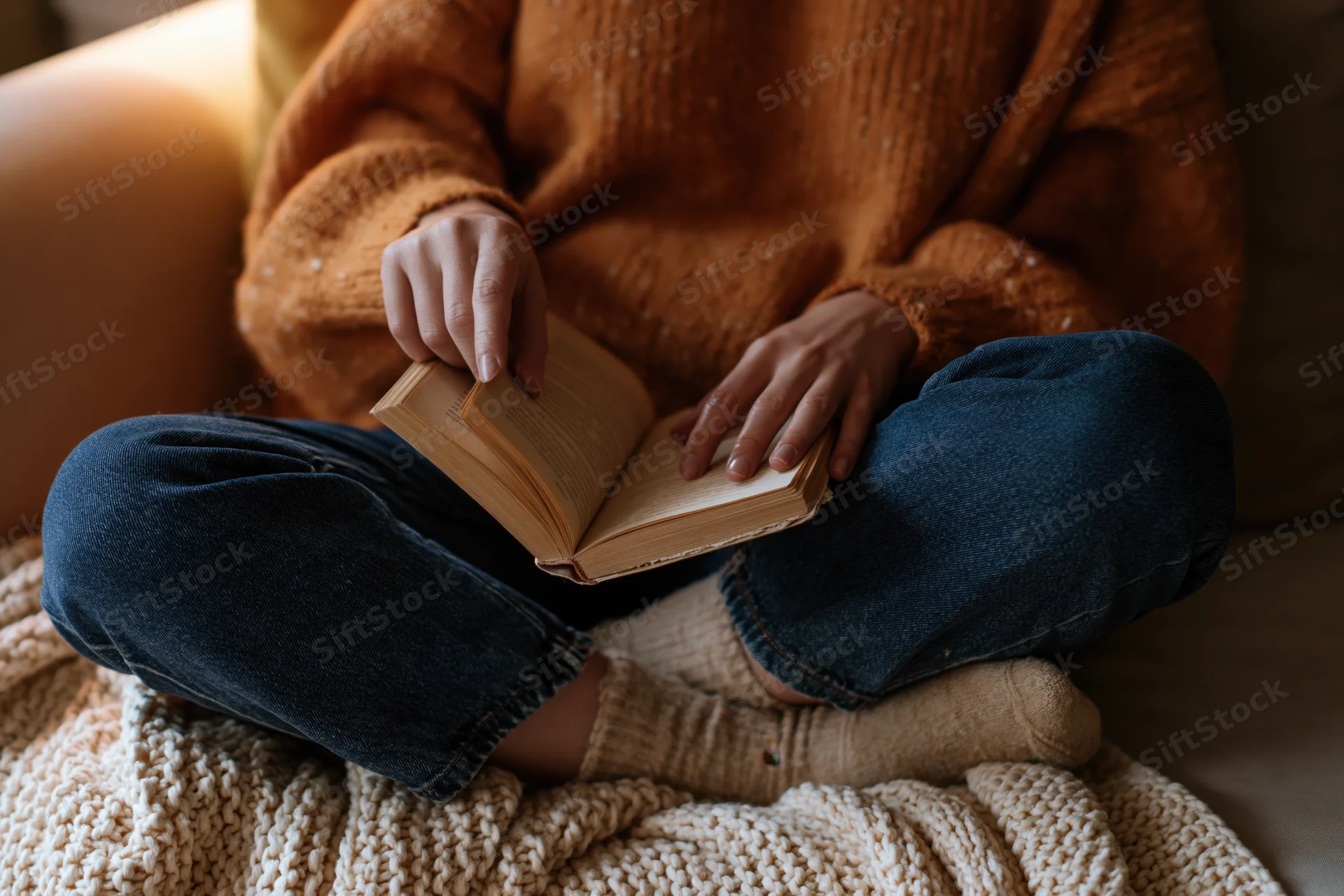 Woman reading a book while sitting cross-legged on a couch covered with a knitted blanket