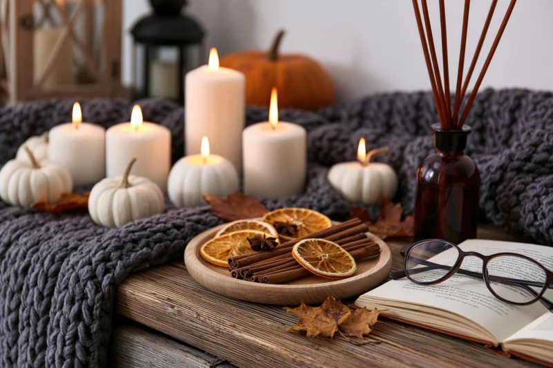 Still life with lit candles, pumpkins, cinnamon sticks, dried oranges, and an open book on a wooden table covered with a knitted blanket