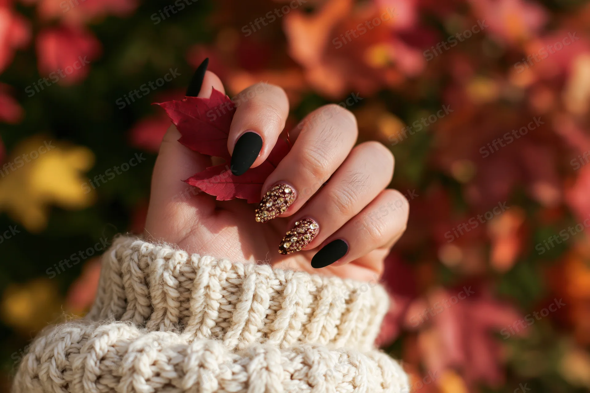 Woman's hand with matte black and glitter nail art holding a red autumn leaf, wearing a beige knitted sweater, outdoors among fall foliage