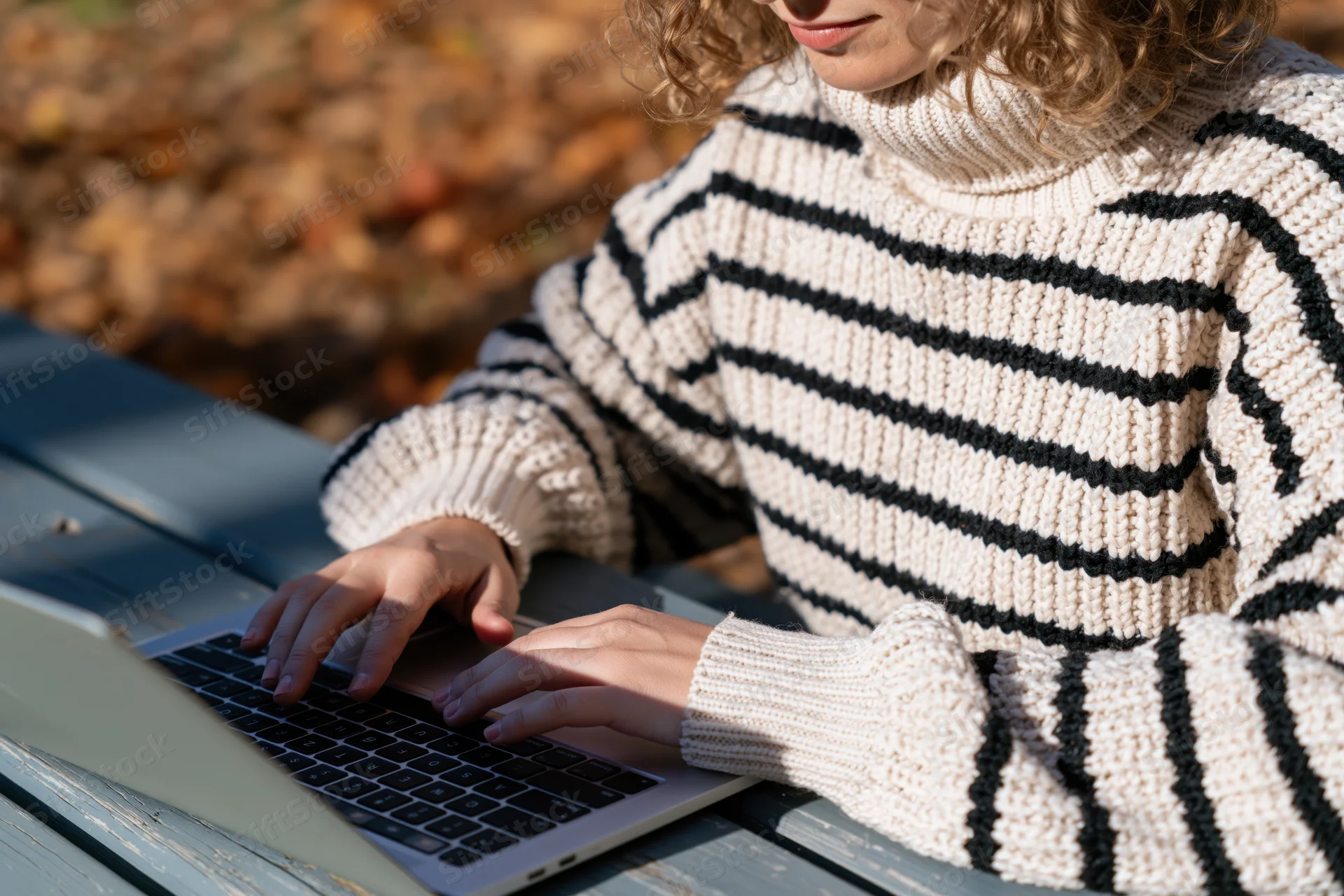 Woman typing on laptop outdoors in autumn foliage wearing striped turtleneck sweater