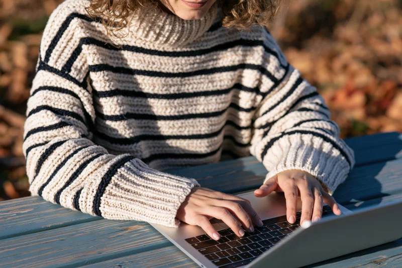 Woman typing on laptop outdoors at wooden table in autumn foliage
