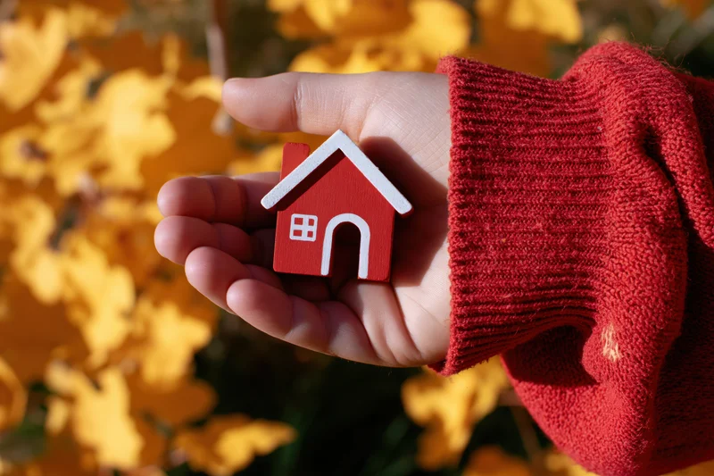 Hand holding a small red wooden house model against a backdrop of yellow autumn leaves