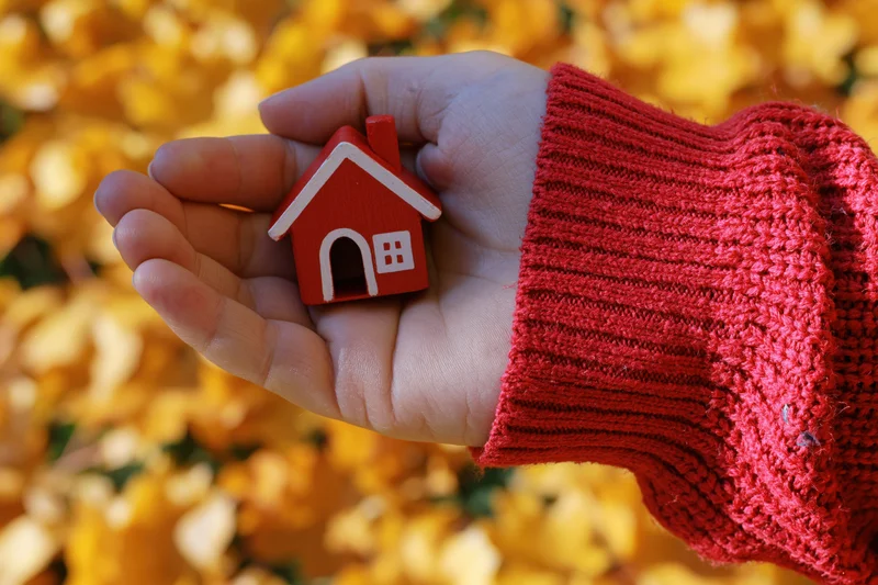 Hand holding small red toy house against blurred autumn leaves background