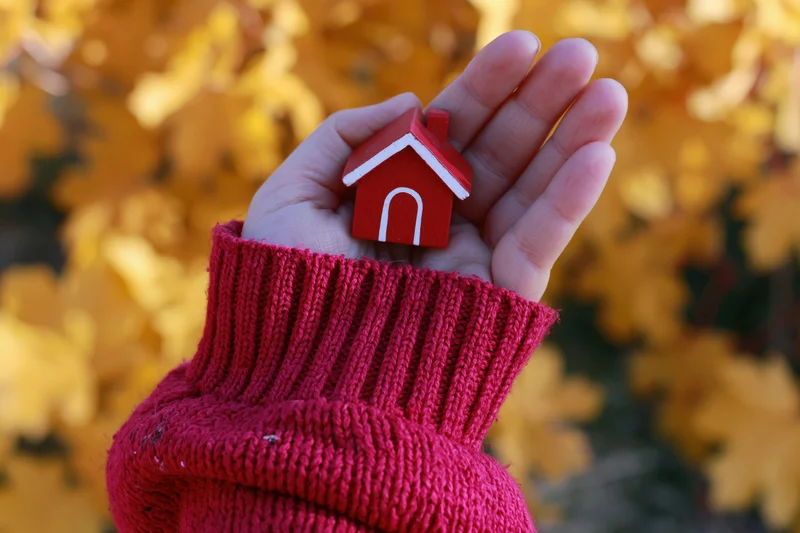 Hand holding a small red house model against a backdrop of yellow autumn leaves