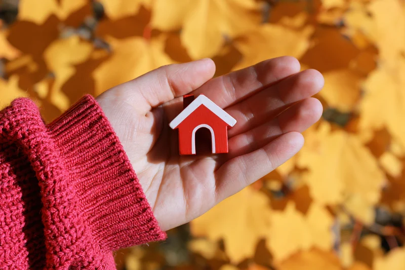 Hand holding small red wooden house model against autumn leaves background