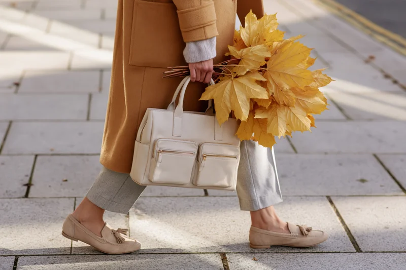 Woman walking on sidewalk carrying bundle of autumn leaves in handbag, wearing coat and loafers