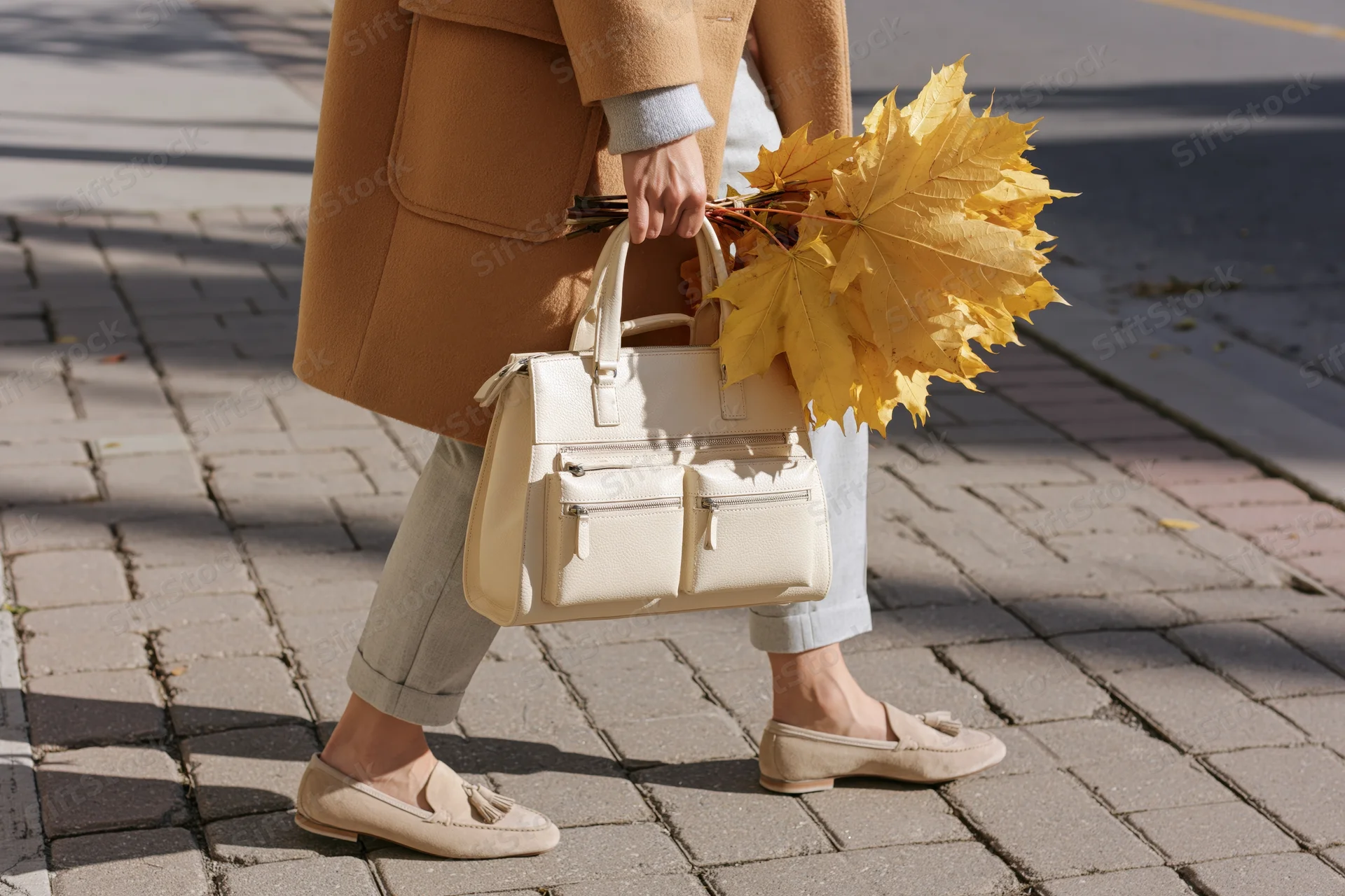 Woman walking on a brick sidewalk carrying a handbag and yellow maple leaves in autumn