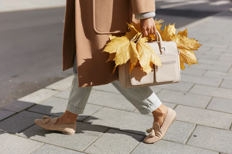 Woman walking on sidewalk carrying handbag with autumn leaves, wearing beige coat and loafers