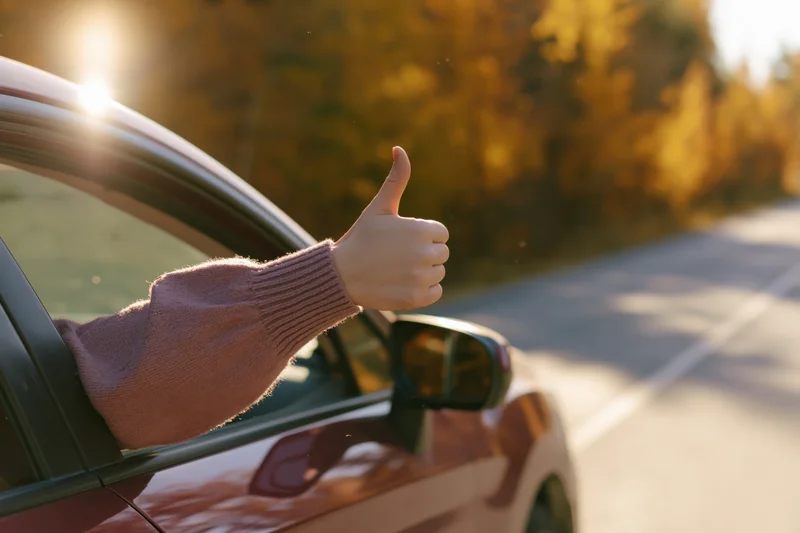 Woman’s hand giving thumbs up from car window on autumn road