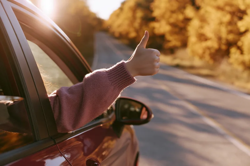 Woman’s arm out of car window giving thumbs up on autumn road