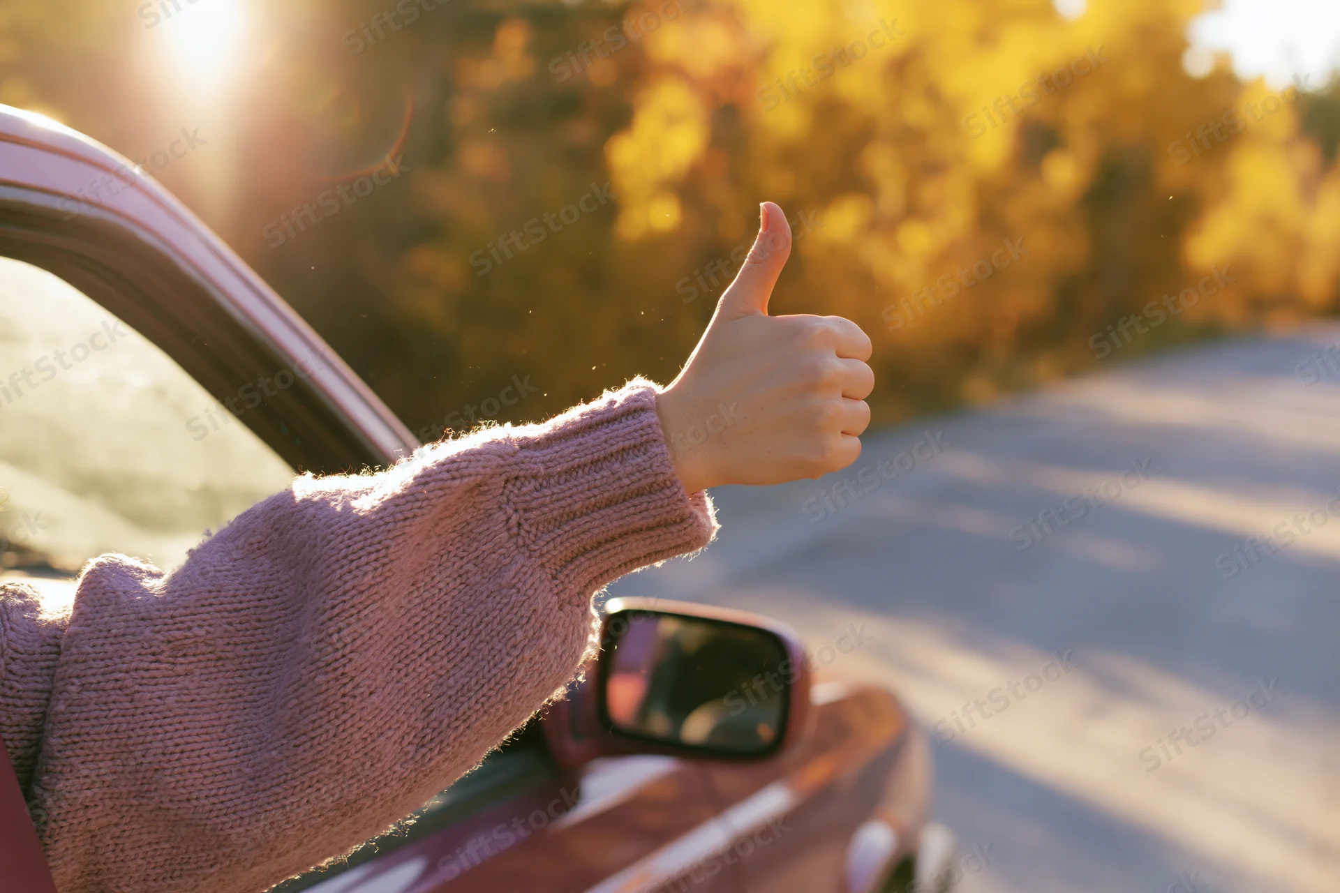 Woman’s arm giving thumbs up from car window on autumn road at sunset