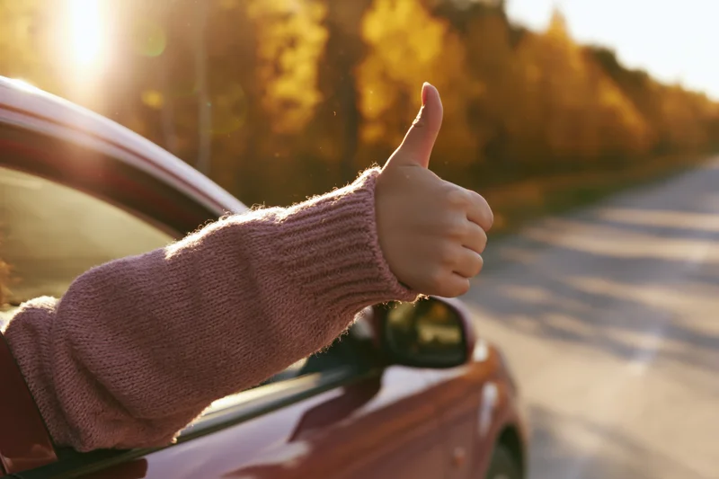 Child’s hand giving thumbs up from car window on autumn road