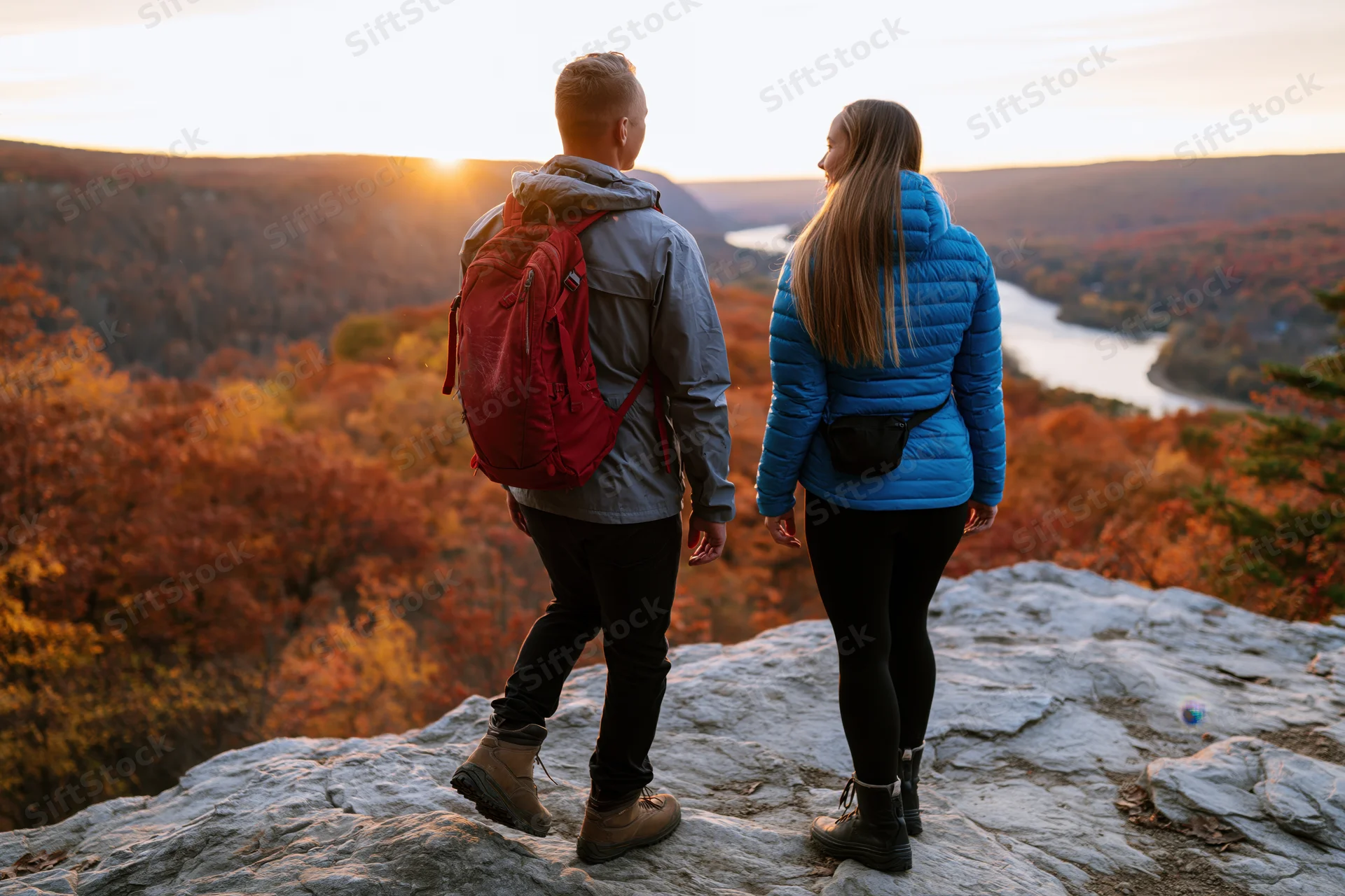 Couple standing on rocky outcrop overlooking autumn forest and river at sunset