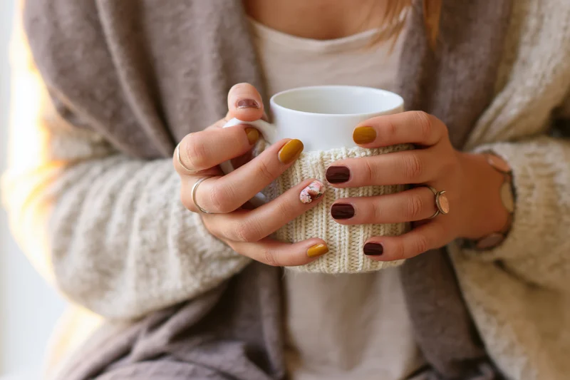Woman holding a white ceramic mug with knitted sleeve, wrapped in a cozy cardigan indoors