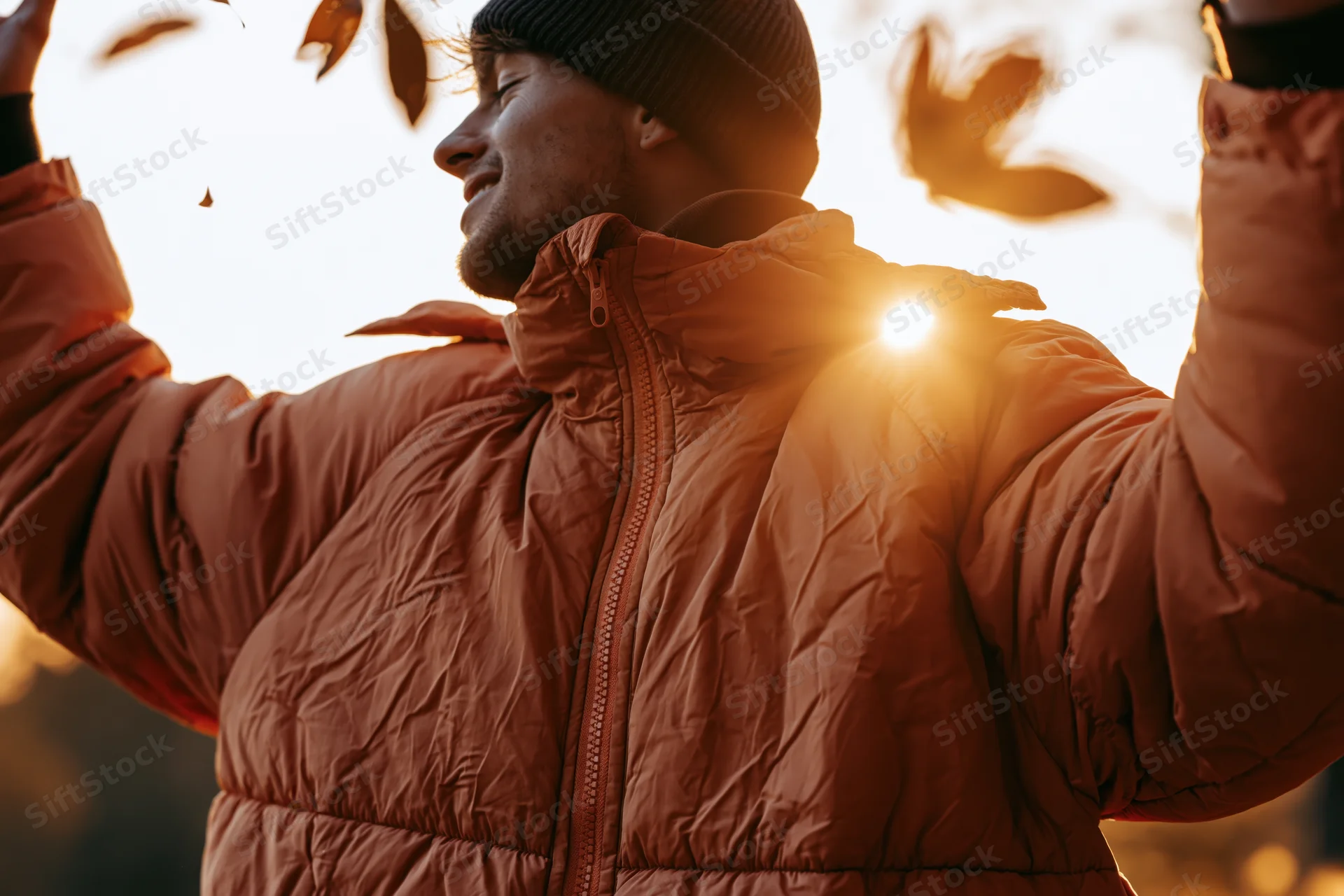Man in puffer jacket with arms raised outdoors during golden hour backlit by sun with falling leaves