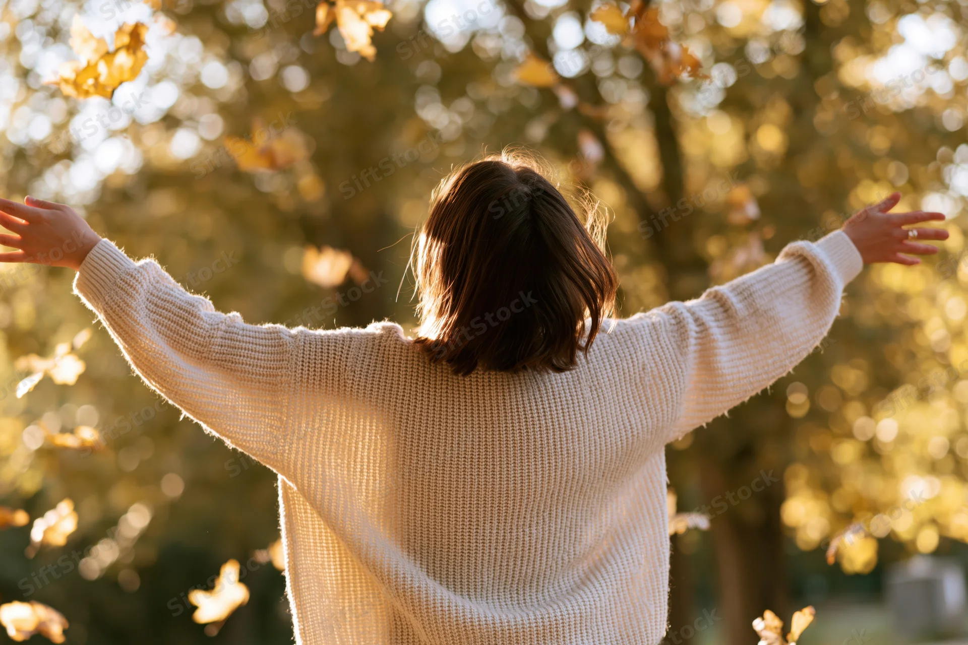 Woman with arms outstretched in autumn park, back to camera, surrounded by golden leaves and sunlight