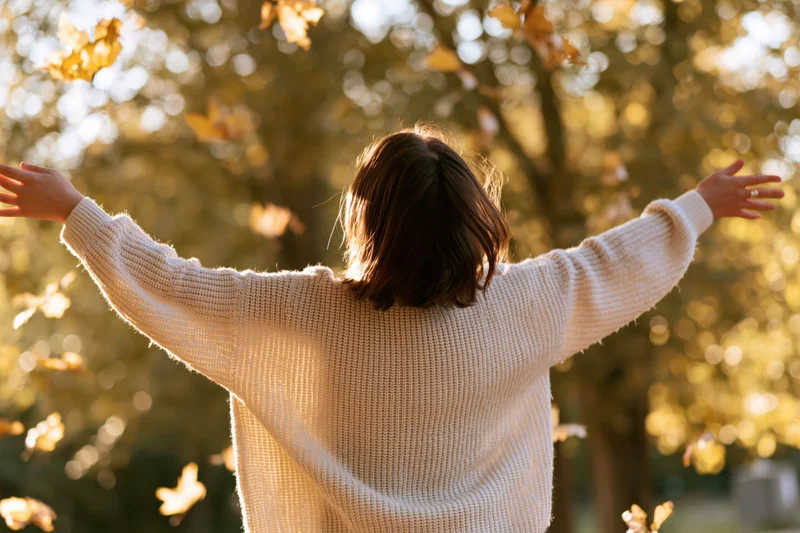 Woman with arms outstretched in autumn park, back to camera, surrounded by golden leaves and sunlight