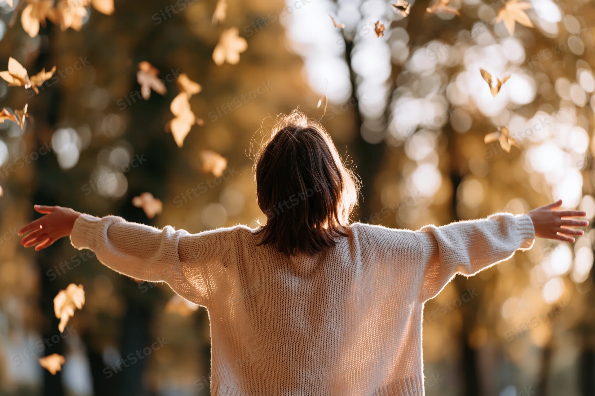 Woman with arms outstretched in autumn forest, back to camera, surrounded by falling leaves in warm sunlight