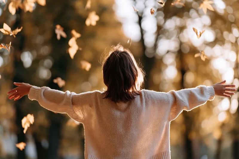 Woman with arms outstretched in autumn forest, back to camera, surrounded by falling leaves in warm sunlight