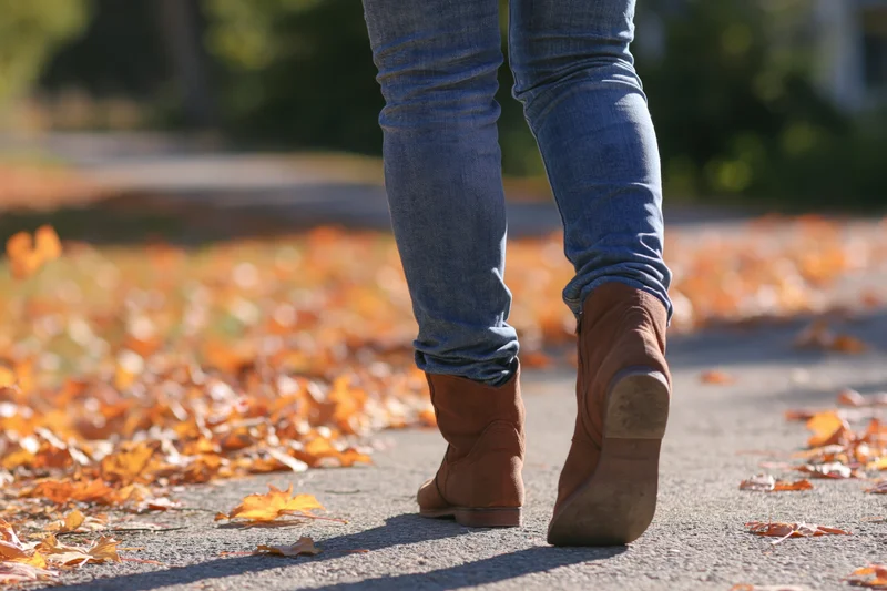 Woman walking away on pavement covered in autumn leaves wearing jeans and brown boots