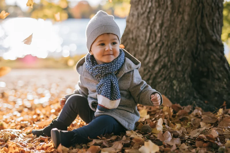Baby sitting on fallen leaves beside a tree in autumn park, smiling and wearing a gray hat and blue scarf