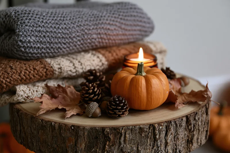 Small pumpkin and lit candle on wooden tree slice with pinecones, leaves, and folded sweaters arranged for autumn decor