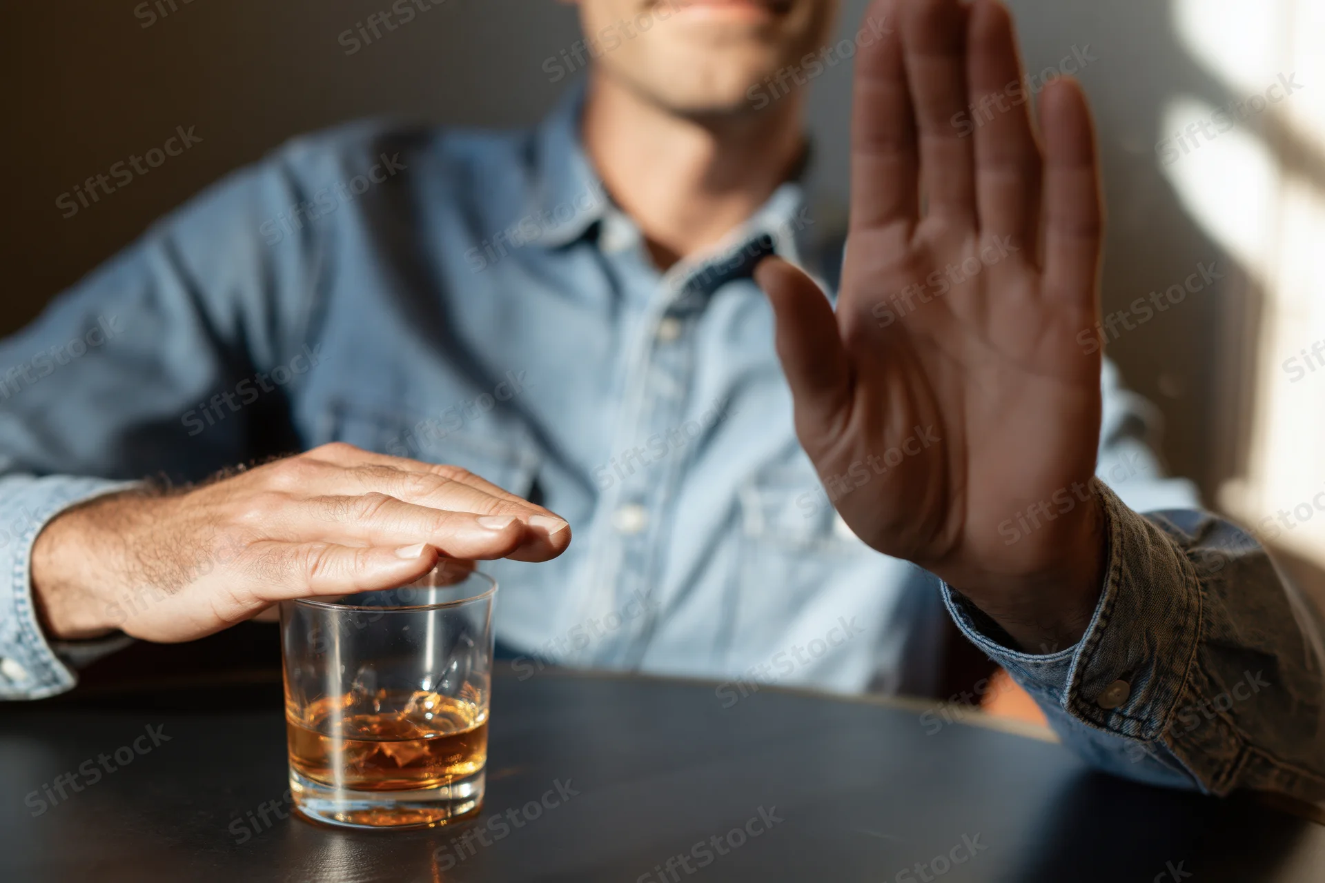 Man raising hand in refusal while holding glass of whiskey on table