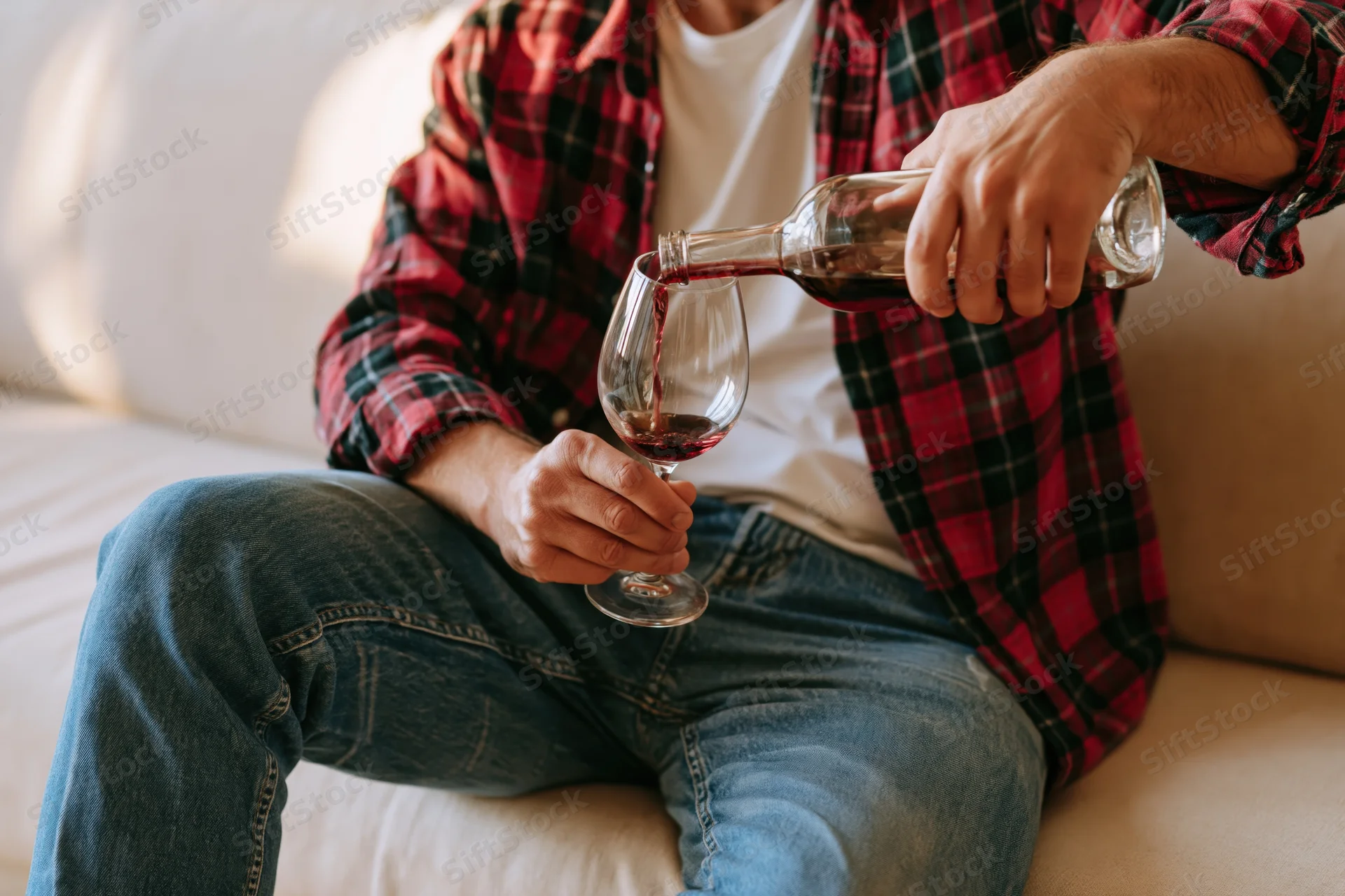 Man pouring red wine into a glass while seated on a beige sofa in a living room