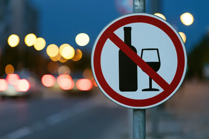 Circular no-alcohol prohibition sign on metal pole by city street at night with blurred vehicle lights in background