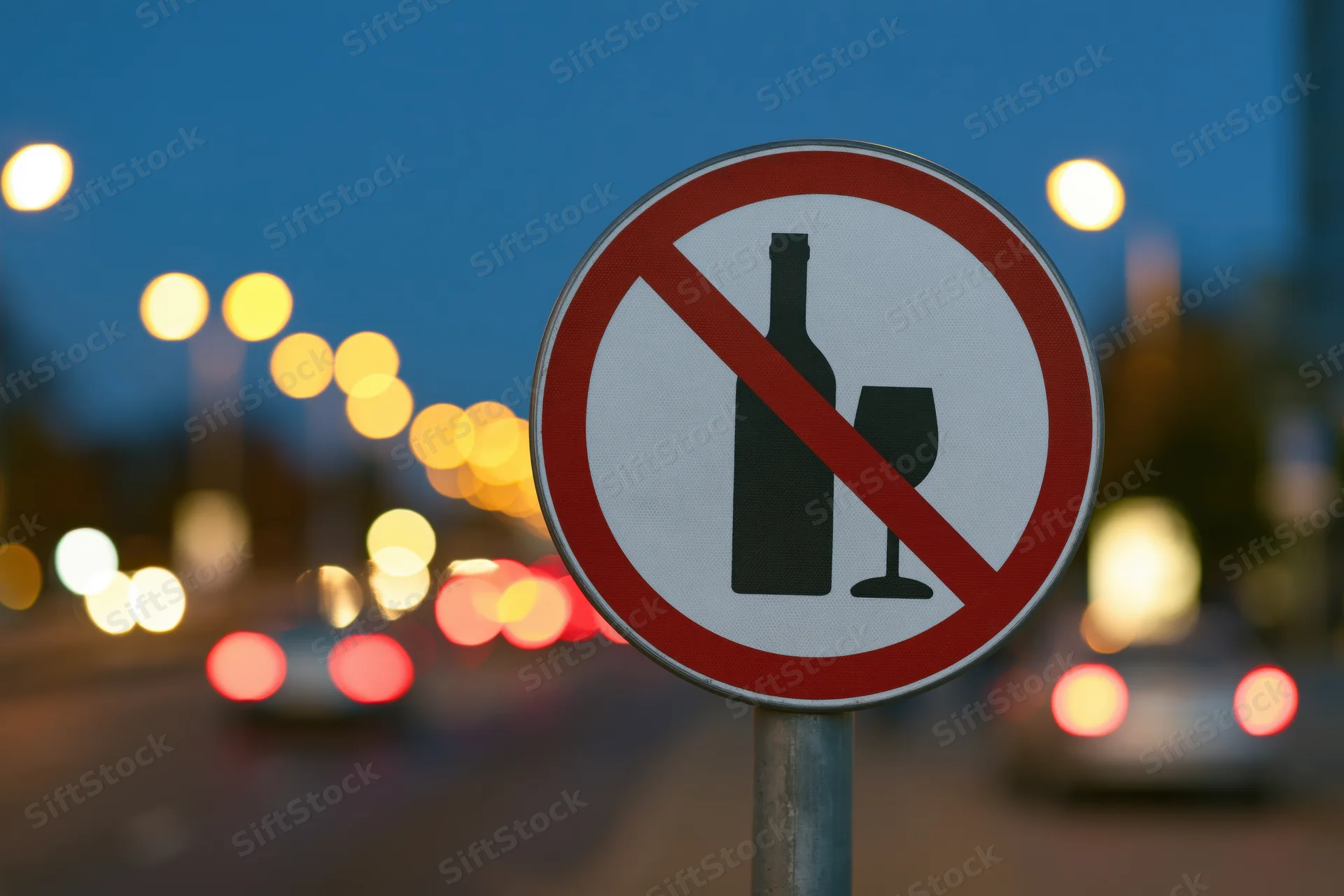 No-alcohol prohibition sign on metal pole by city street at twilight with blurred car lights in background