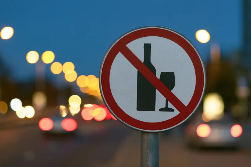 No-alcohol prohibition sign on metal pole by city street at twilight with blurred car lights in background