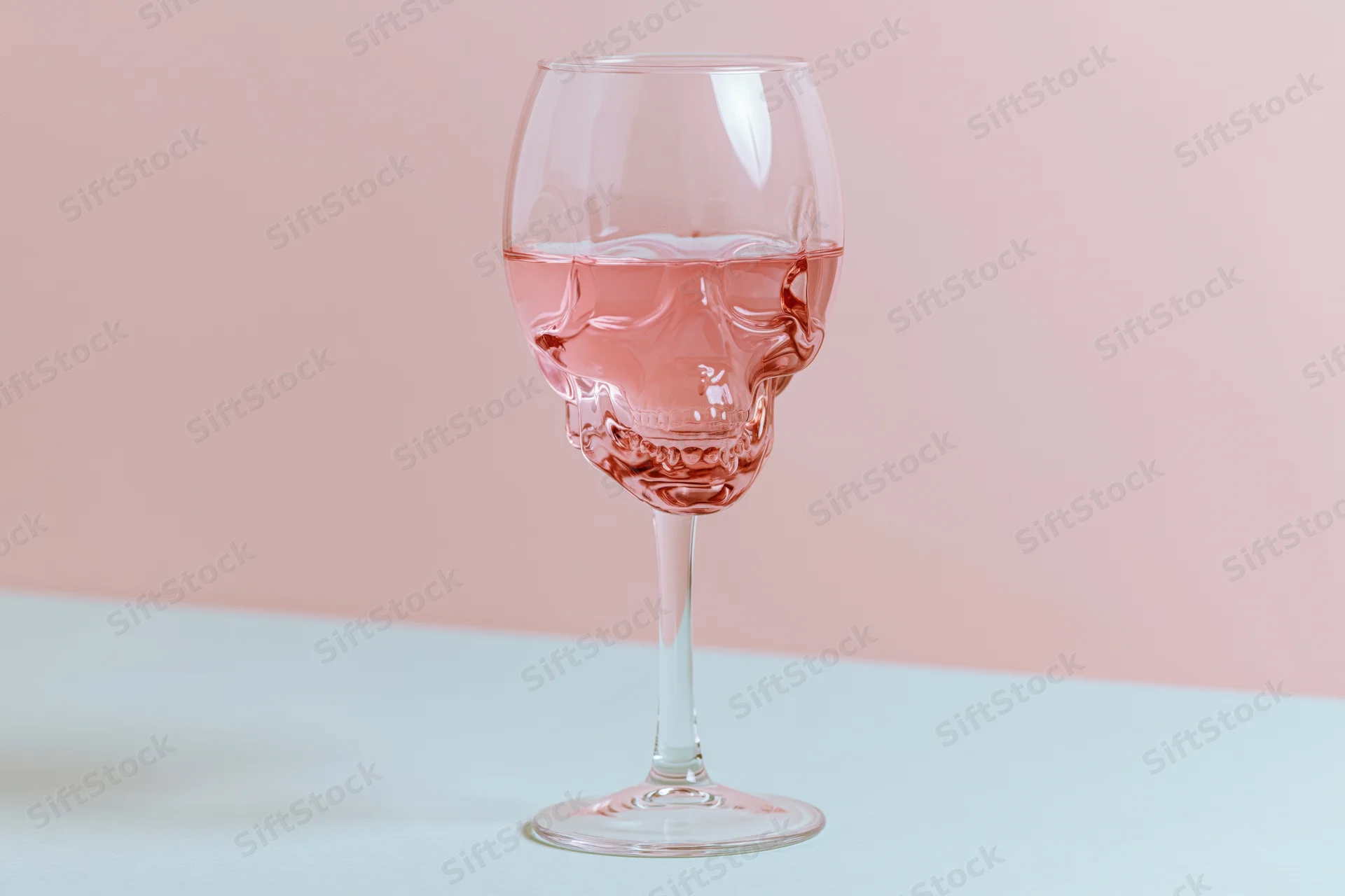 Skull-shaped wineglass filled halfway with pink liquid on a pastel surface