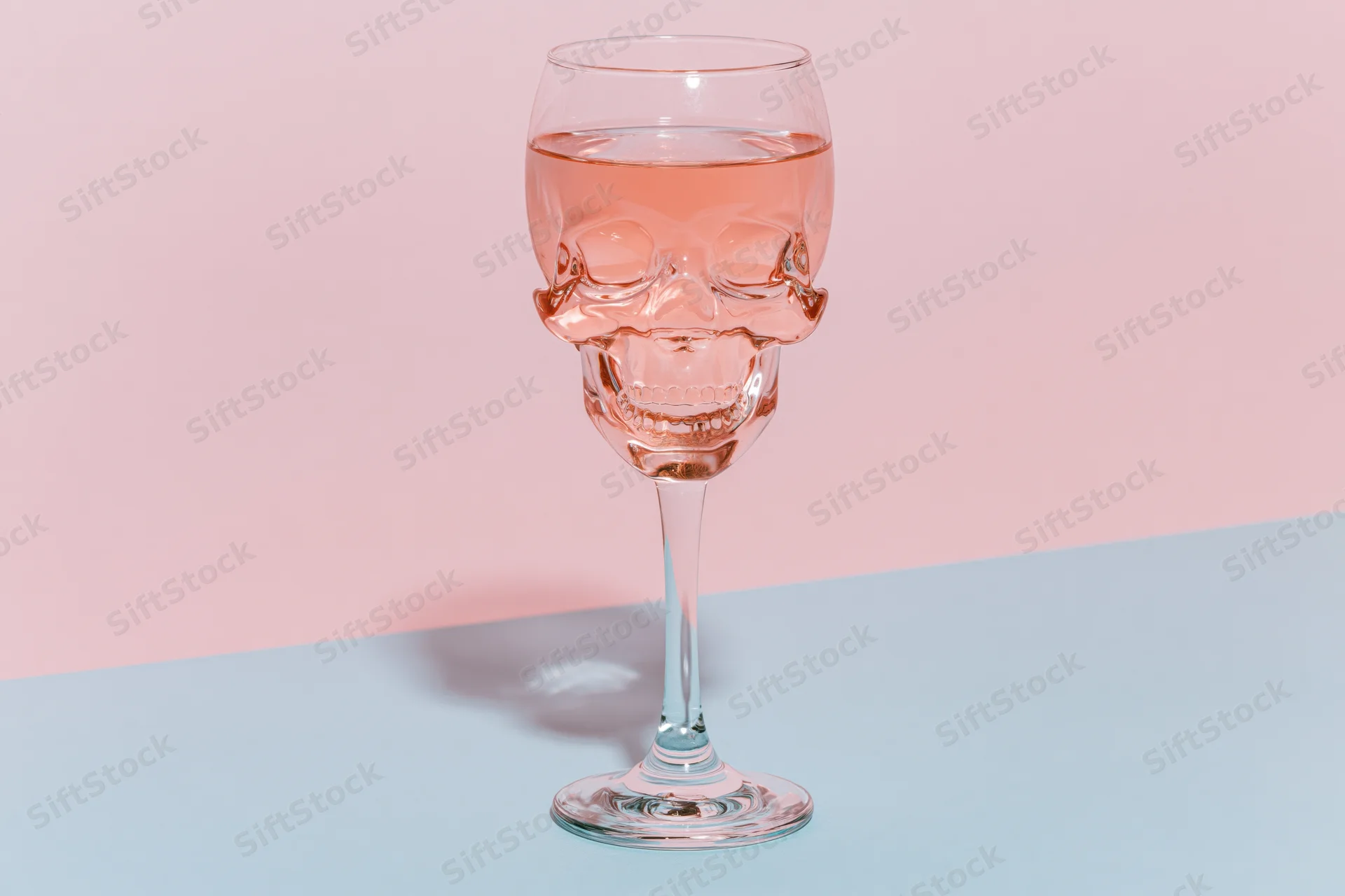 Skull-shaped wine glass filled halfway with pink liquid on a pastel blue and pink surface