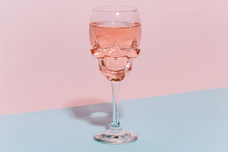 Skull-shaped wine glass filled halfway with pink liquid on a pastel blue and pink surface