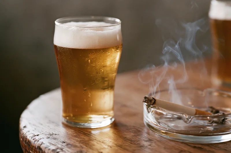 Pint glass of golden lager with frothy head and smoking cigarette in ashtray on wooden table