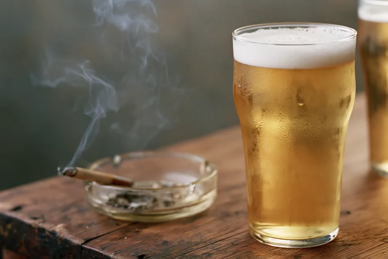 Full pint glass of pale golden lager with frothy head and smoking cigarette in ashtray on wooden table