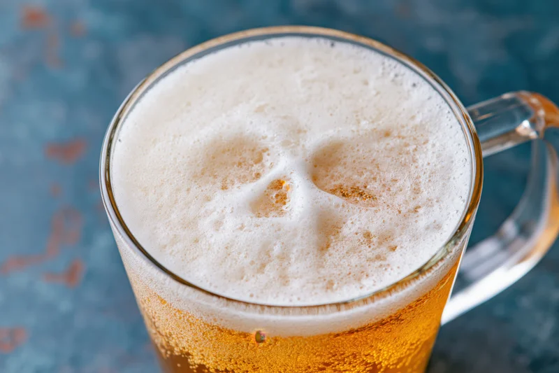 Glass beer mug filled with golden lager and thick white foam shaped like a skull on a blue textured surface