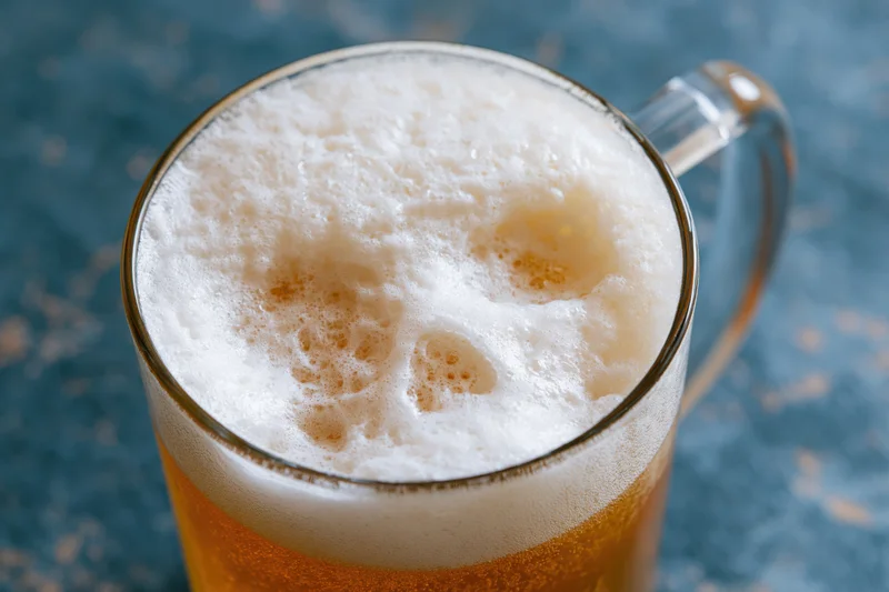 Glass beer mug filled with golden lager and thick white foam on blue surface