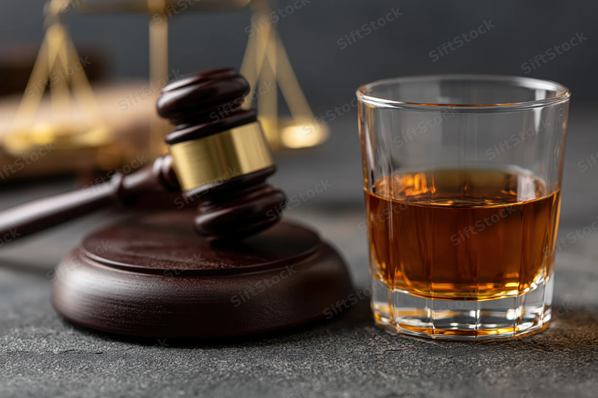 Glass of amber whiskey beside a wooden judge's gavel on a dark surface with scales of justice in background