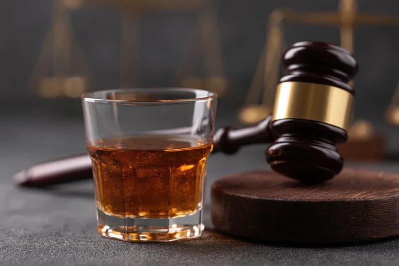Glass of amber whiskey beside a wooden judge's gavel on a dark surface with scales of justice in the background