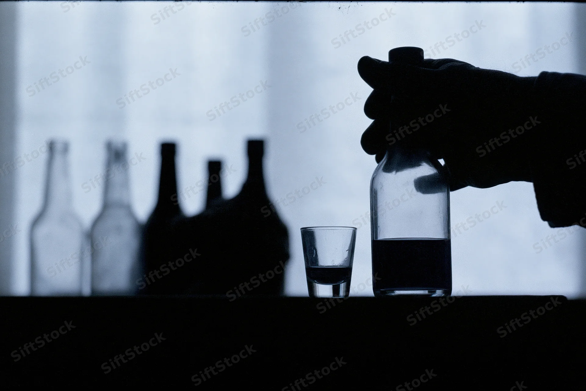 Hand gripping a liquor bottle next to a shot glass on a table in silhouette