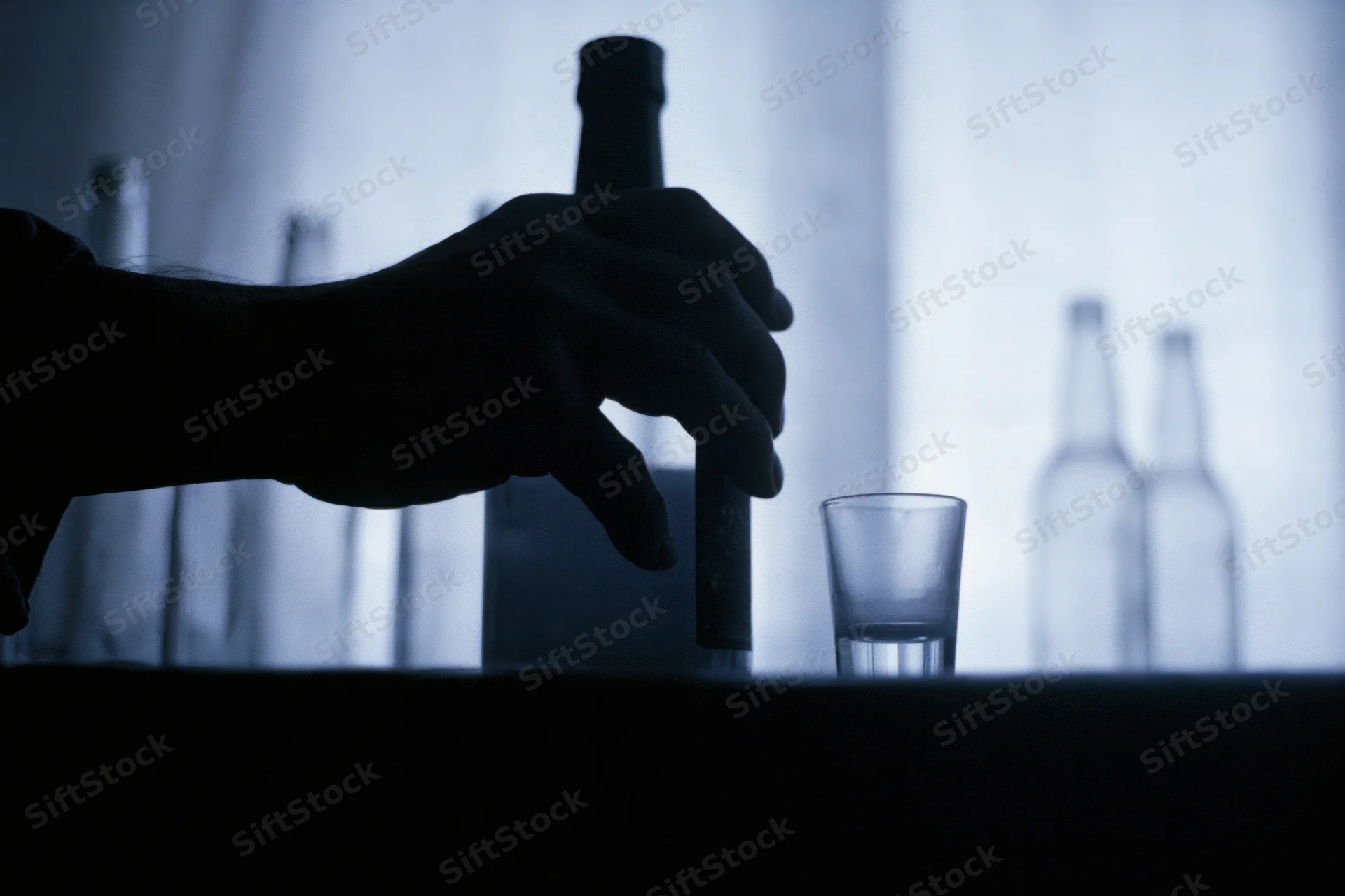 Hand gripping a liquor bottle next to a shot glass on a bar counter in dim blue light