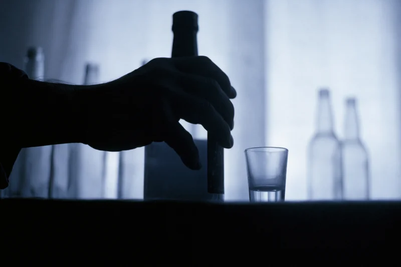 Hand gripping a liquor bottle next to a shot glass on a bar counter in dim blue light