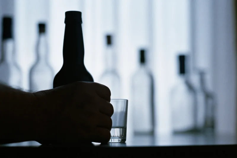 Hand gripping a liquor bottle next to a shot glass on a table in dim light