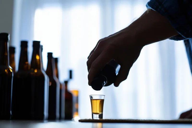 Hand pouring beer into a shot glass on a table with bottles in the background