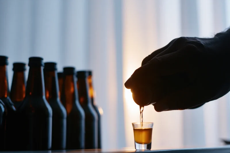 Hand pouring amber liquid into shot glass with beer bottles in silhouette against glowing curtain backdrop