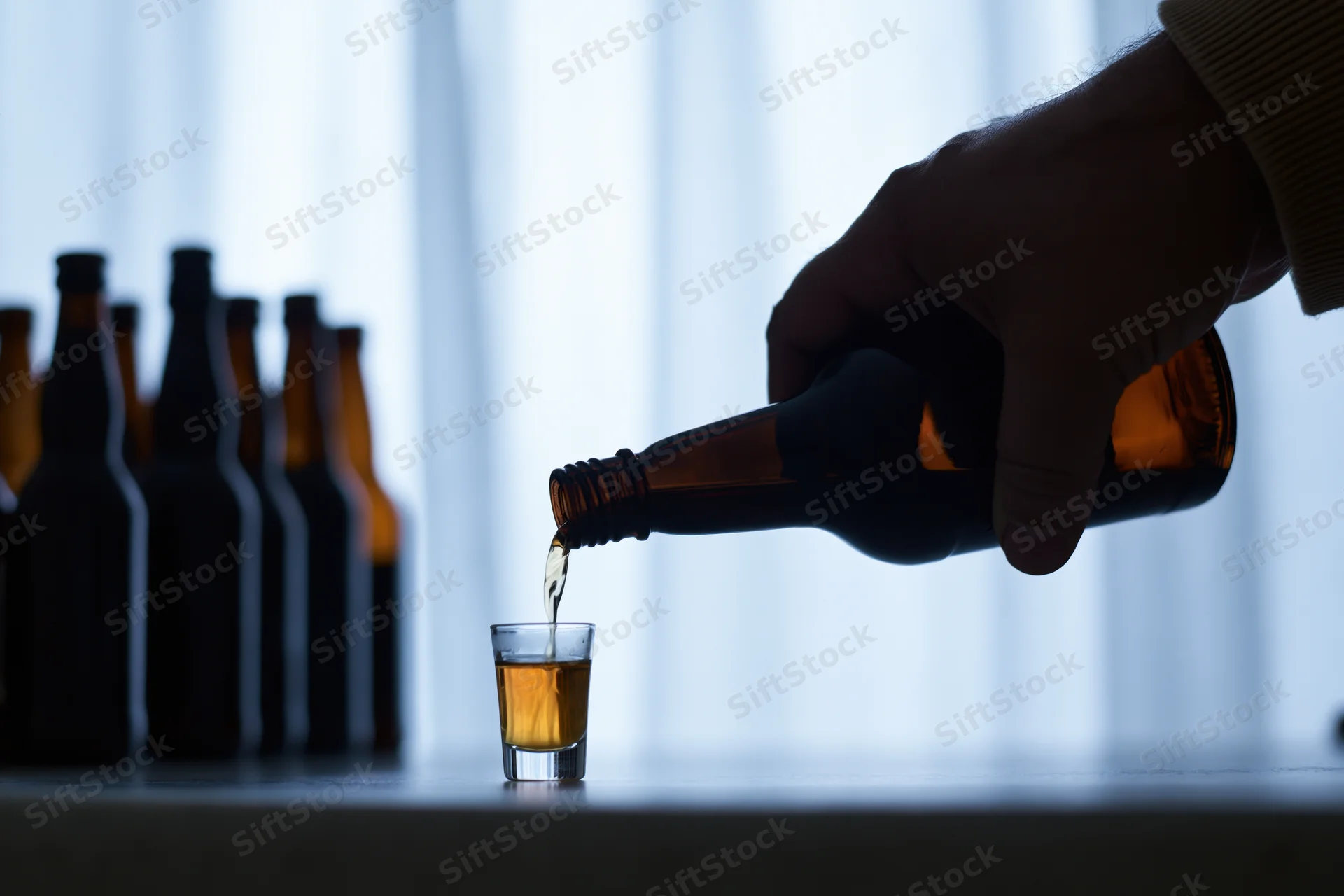 Hand pouring amber liquid from a bottle into a shot glass on a table against a glowing curtain backdrop