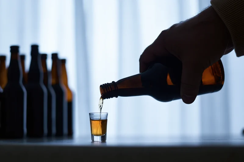 Hand pouring amber liquid from a bottle into a shot glass on a table against a glowing curtain backdrop