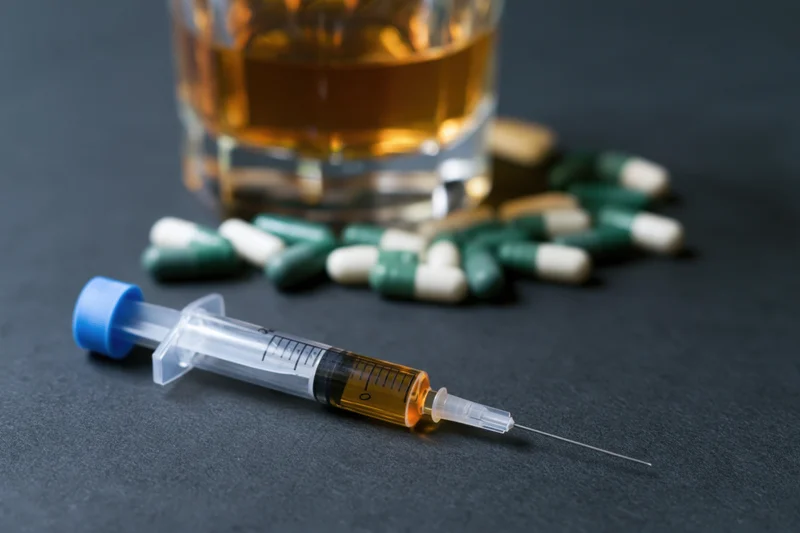 Syringe with blue cap lying on dark matte surface beside glass of amber liquid and scattered pills