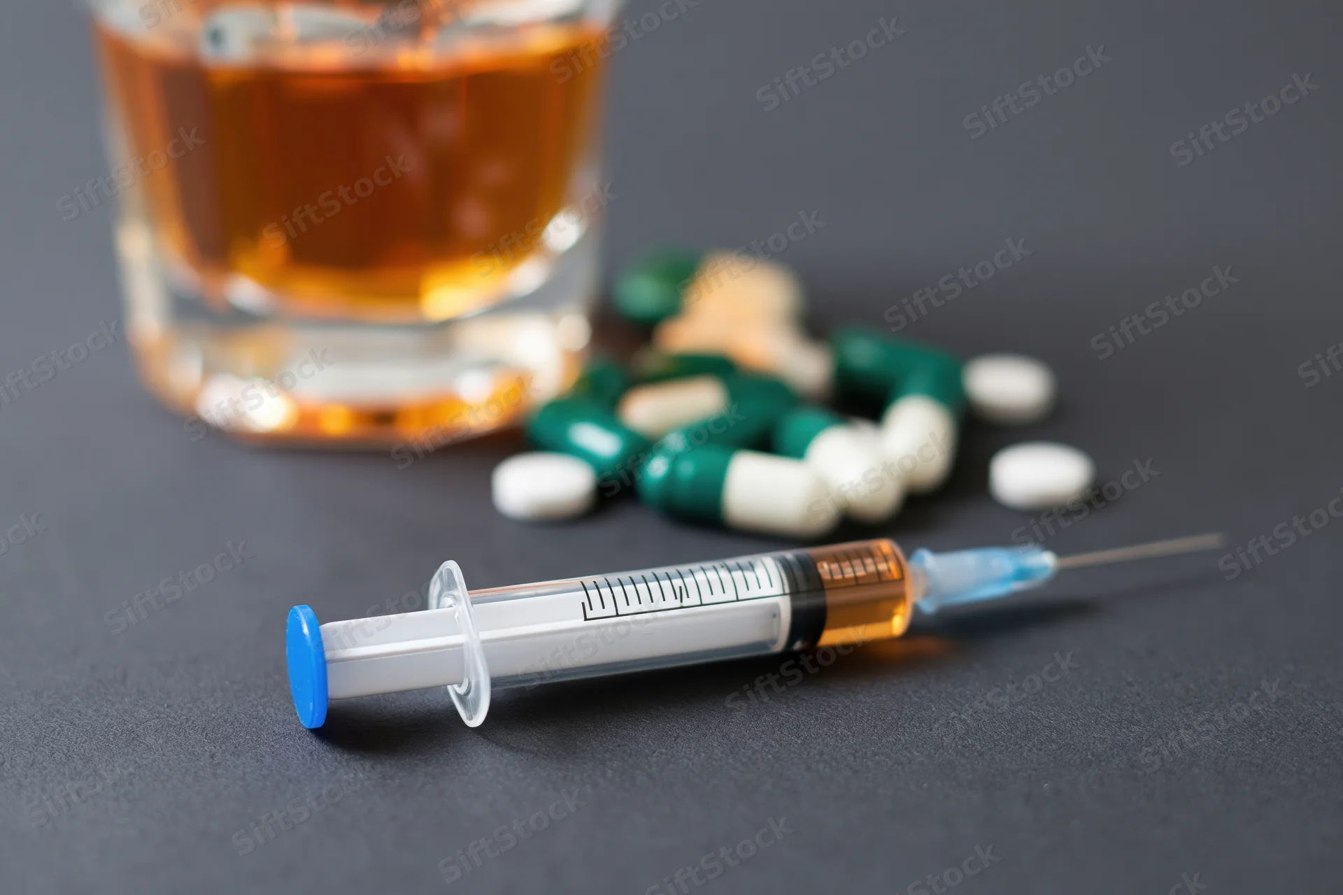 Syringe with blue cap lying on dark matte surface beside glass of amber liquid and scattered pills